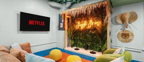 Una habitación con un cartel de Netflix, un muro de paja y diferentes opciones de asiento, incluyendo una silla verde y un sofá.