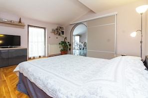 Ein Schlafzimmer mit einem großen Bett, einem Fernseher, einer Topfpflanze, einer Schiebetür und Blick auf einen Balkon.