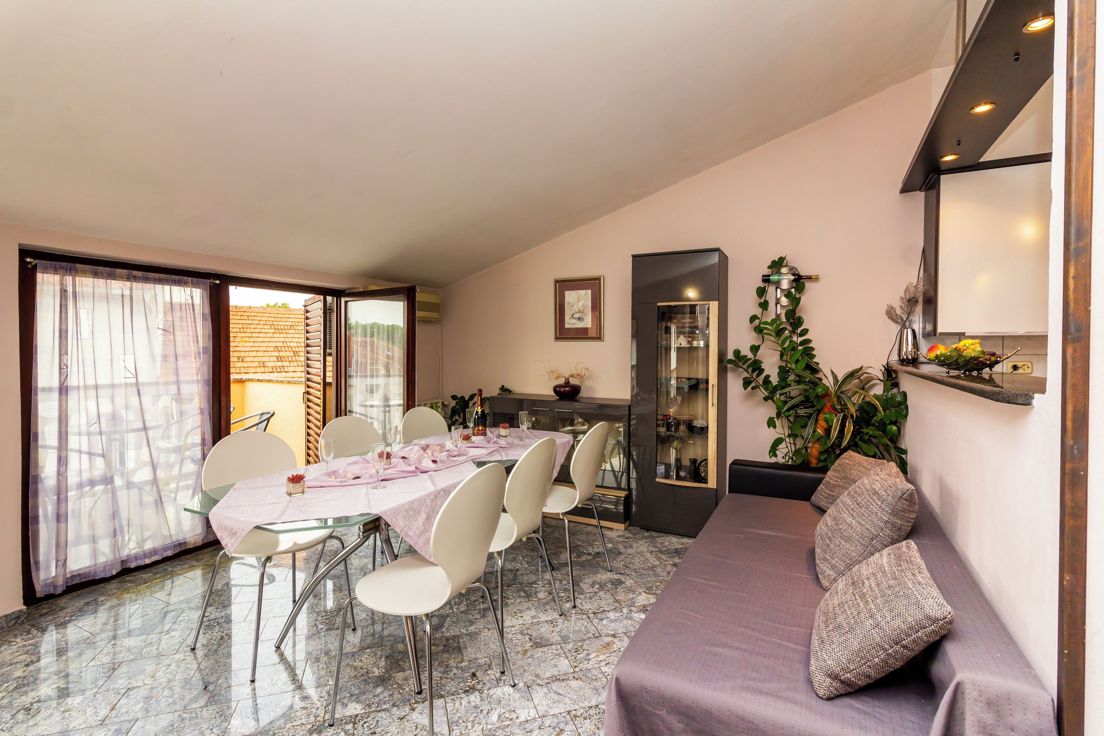 A dining area with a table set for four, a sofa with cushions, a plant, and a kitchen area in the background.