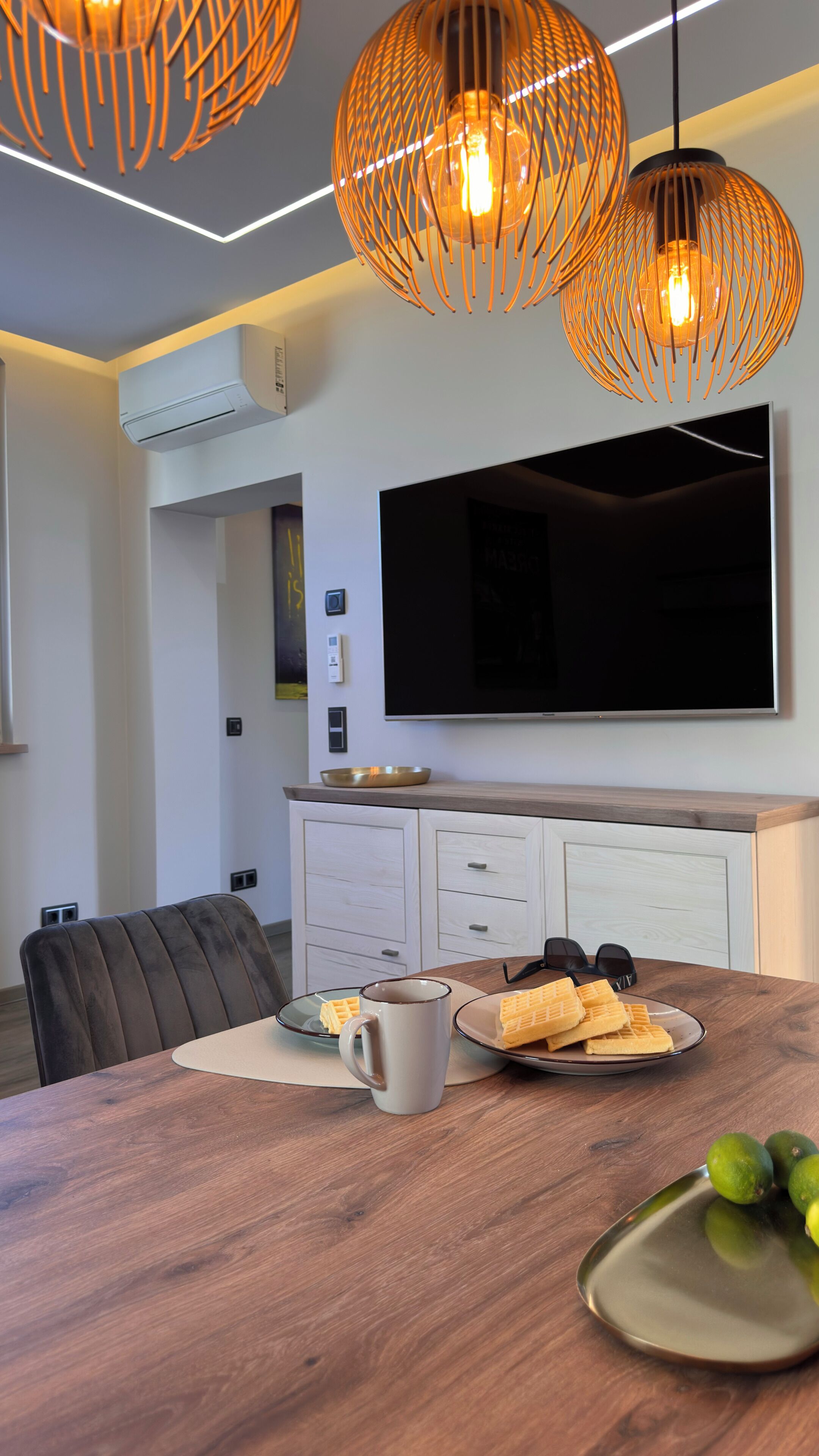 A modern kitchen with a flat-screen TV, wooden cabinets, and a dining table with waffles and a mug.