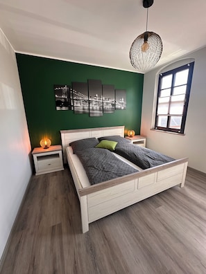Ein Schlafzimmer mit Bett, Nachttischen, einer Hängeleuchte, einem Fenster und einer grünen Wand mit Stadtbild-Kunstwerk.