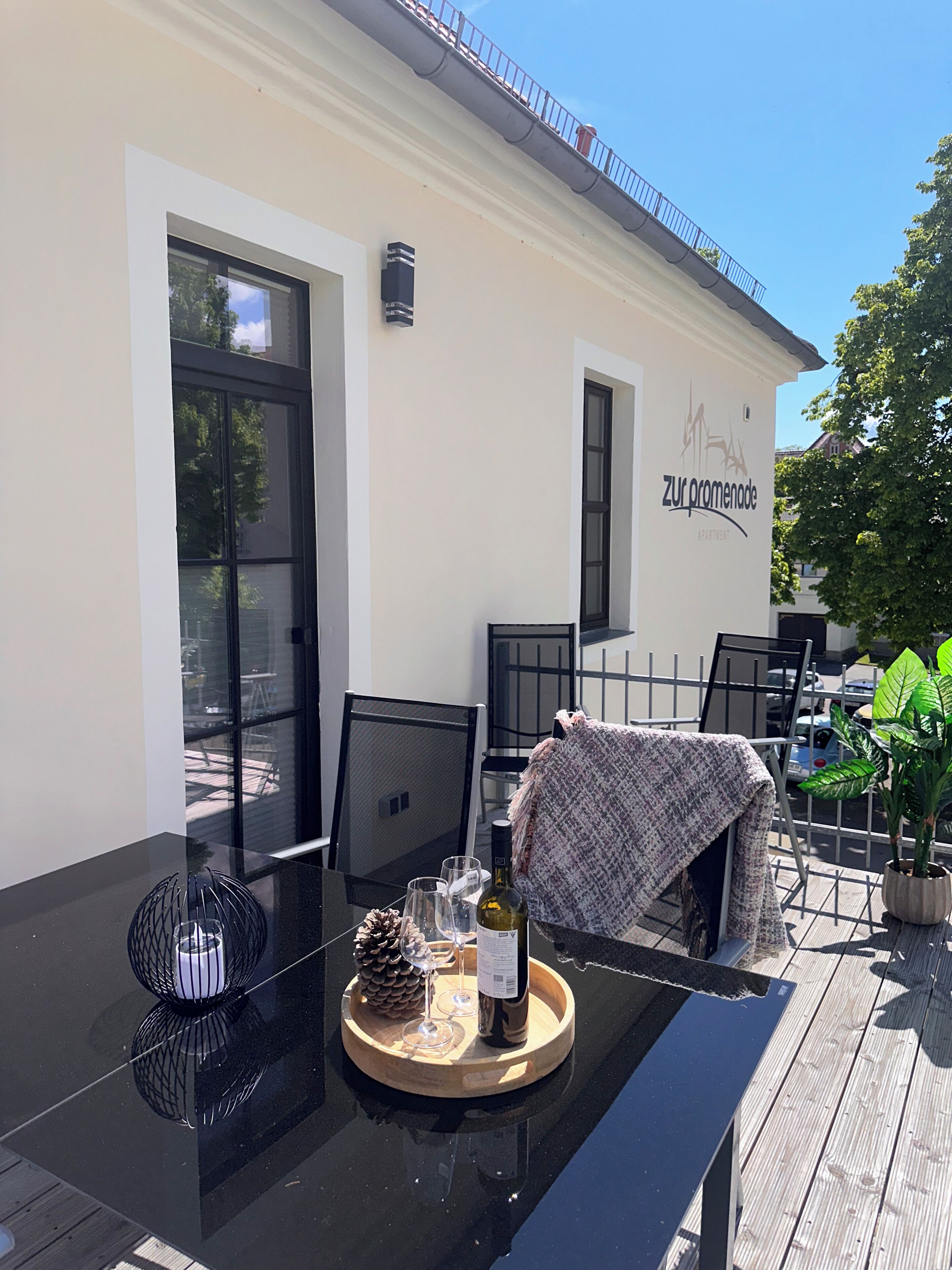 An outdoor dining area with a table set for two, chairs, a potted plant, and a building with a sign reading 'zur promenade apartments'.