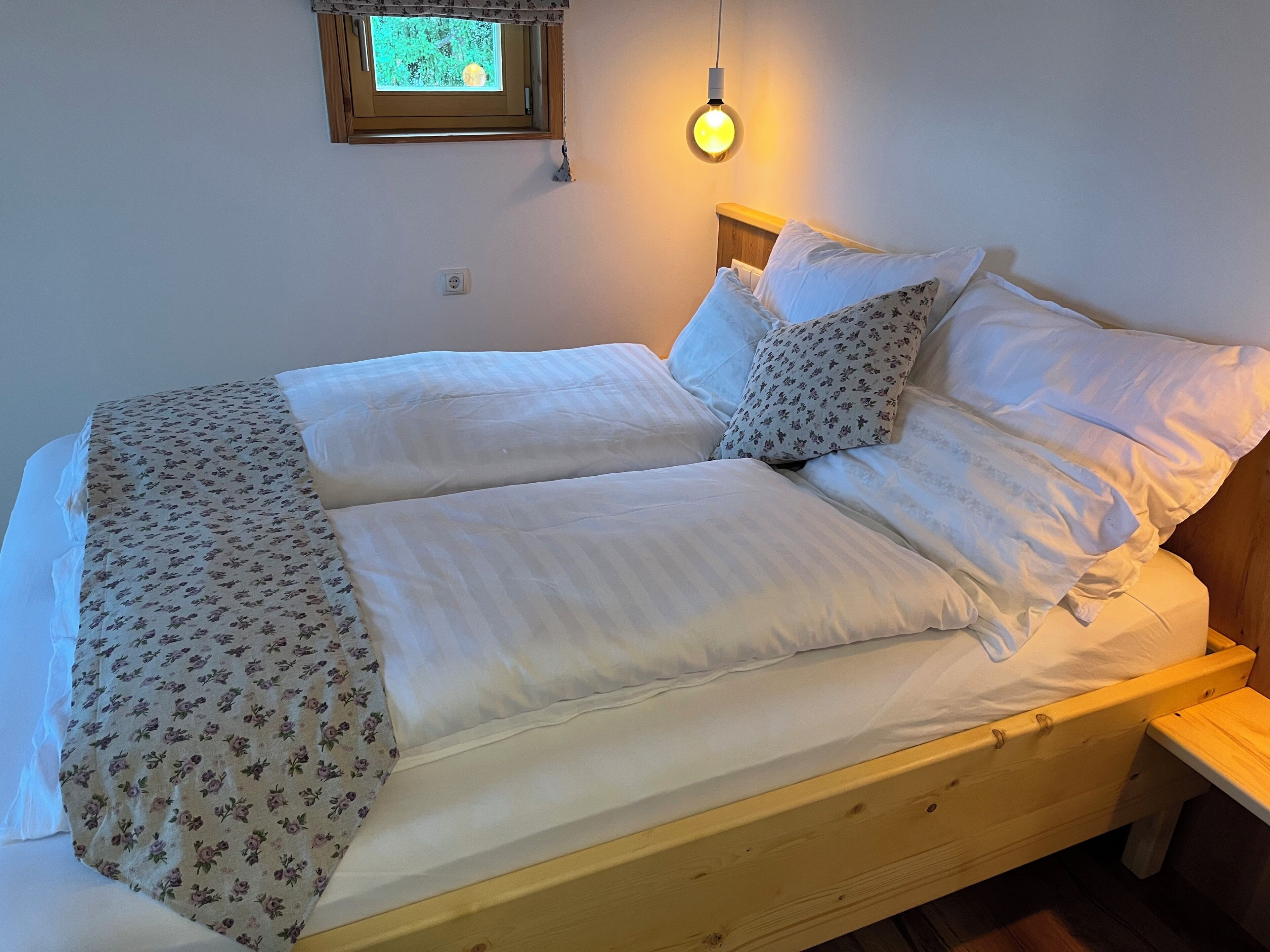 A double bed with floral-patterned bedding, a bedside table, a nightstand, a window with a view, and a hanging light fixture.