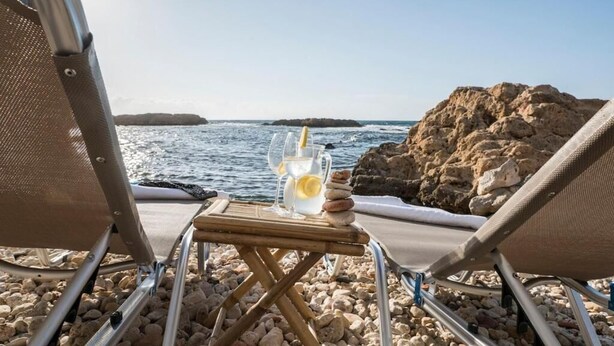 Uitzicht op het strand met een tafel waarop een drankje staat, rotsen, twee strandstoelen en een rotsachtige kustlijn.