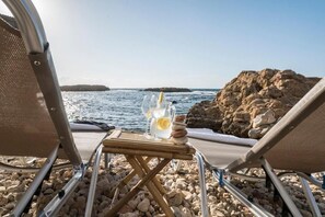 A beachside view with a table holding a drink and rocks, two beach chairs, and a rocky shoreline. - Kito Transformed House - Sea View (Chania)