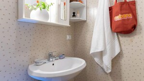 Ein Badezimmer mit einem weißen Waschbecken, einem Spiegel darüber und einem Handtuch an der Wand. Eine rote Tasche hängt am Handtuchhalter.