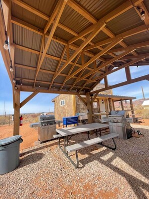 A covered outdoor area with a barbecue grill, picnic table, and wooden structure in the background.