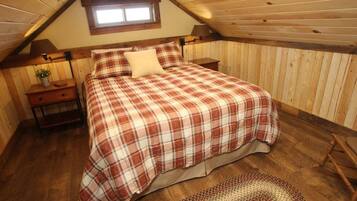 Un dormitorio con techo inclinado, una cama con cubrecama escocés, una mesita de noche con lámpara y un ventanal pequeño.