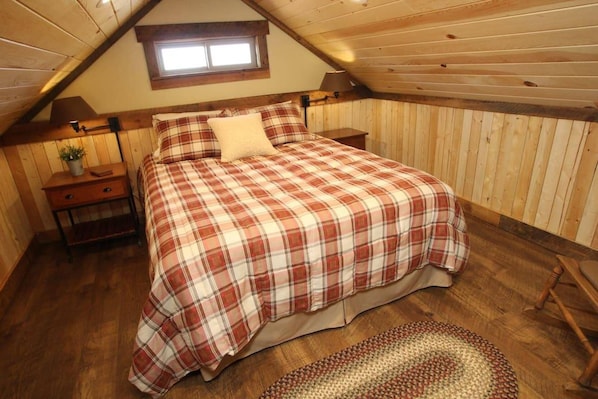 Un dormitorio con techo inclinado, una cama con cubrecama escocés, una mesita de noche con lámpara y un ventanal pequeño.