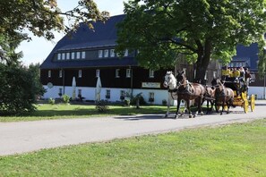 A building with a dark roof and white walls, a horse-drawn carriage with passengers, and a tree-lined driveway. - Waldgasthof Bad Einsiedel (Seiffen/Erzgebirge)