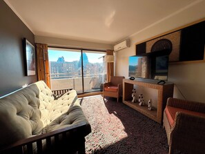 Ein Hotelzimmer mit großem Fenster, einer Couch, einem Sessel, einem Fernseher und einem kleinen Tisch mit Figuren.