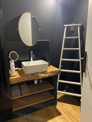 Une salle de bain avec un lavabo blanc, un miroir rond, un porte-serviettes et une étagère en bois.