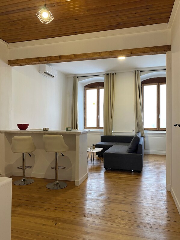 A modern living space with a kitchen area, bar stools, a sofa, and large windows.