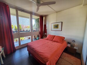 Ein Schlafzimmer mit Bett, Deckenventilator, Fenster mit Vorhängen, einem gerahmten Bild an der Wand und Blick auf den Balkon.