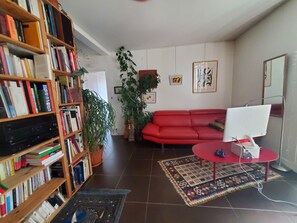 Ein Wohnzimmer mit einem roten Sofa, einem Holzbücherregal, einem Desktop-Computer und einem gemusterten Teppich.