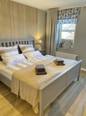 A neatly made bed with a striped bedspread and pillows, a bedside table with a lamp, a window with curtains, and a radiator. - Large Baltic Sea Nest (Boltenhagen)