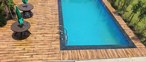 Una piscina rectangular con un muelle de madera alrededor, una escalera metálica y dos sombrillas verdes.