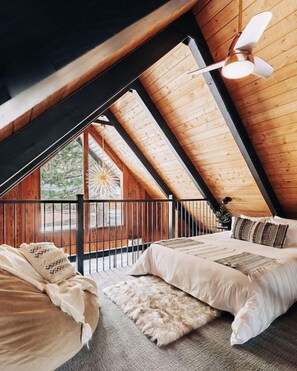 A bedroom with a sloped ceiling, a bed with white bedding, a hanging light fixture, and a view of the outdoors. - Firefly Cabin with Mountain View (Big Bear Lake)