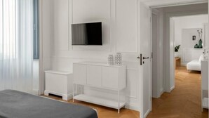 Un dormitorio moderno con una cama, un televisor, un mueble blanco y un pasillo que da a una sala de estar.