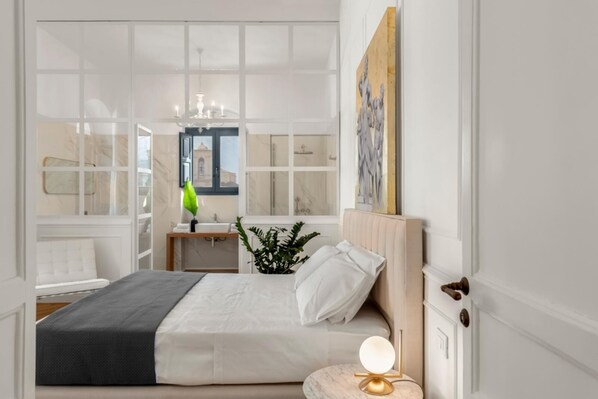Ein Schlafzimmer mit Bett, Nachttisch, Lampe, Topfpflanze und Blick ins Badezimmer durch ein Fenster.