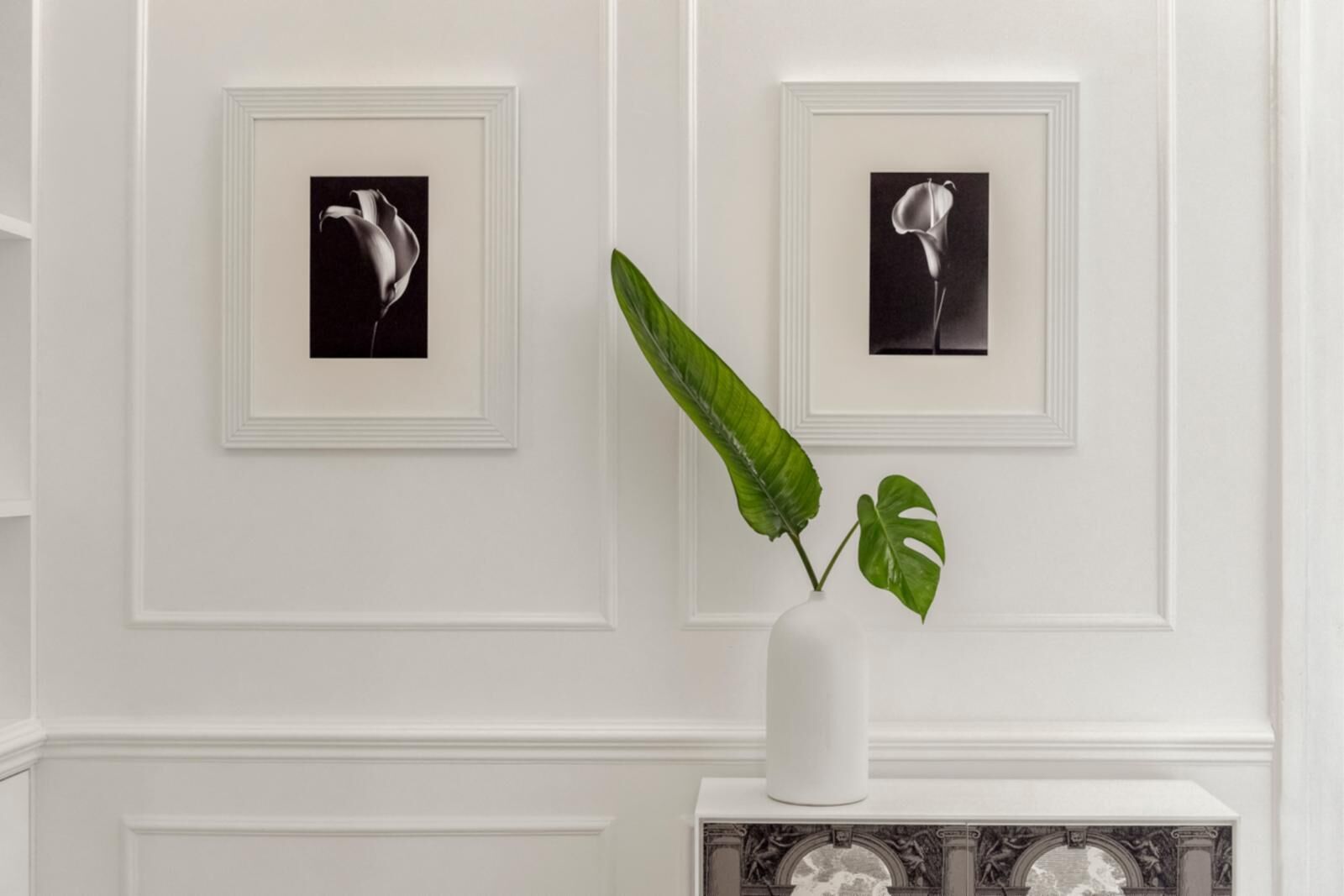 Een witte muur met twee ingelijste zwart-witfoto's van bloemen, een witte vaas met een plant, en een decoratieve voet.