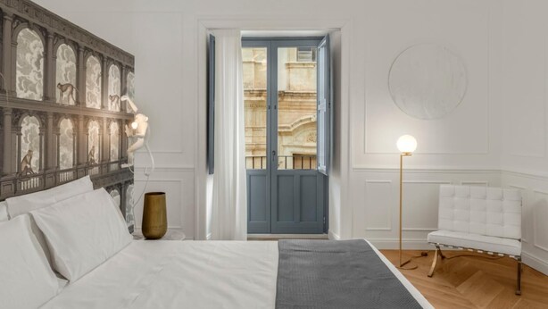 Un dormitorio con cama, mesita de noche, un jarrón, lámpara y vista a un edificio a través de una puerta.