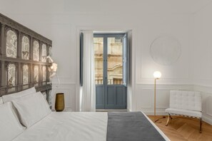 Un dormitori amb un llit, una taula de nit, un vas, una làmpada i vista d'un edifici a través d'una porta.