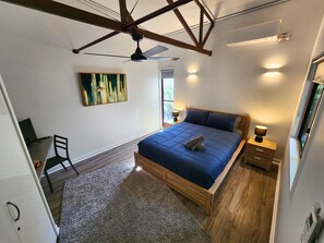 A bedroom with a bed, a desk, a chair, a ceiling fan, a painting, and a window.