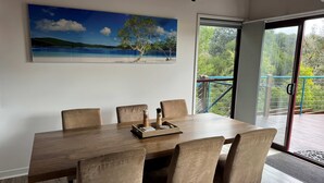 Un comedor con una mesa de madera y seis sillas, un cuadro de una playa y vistas a un balcón con vegetación.