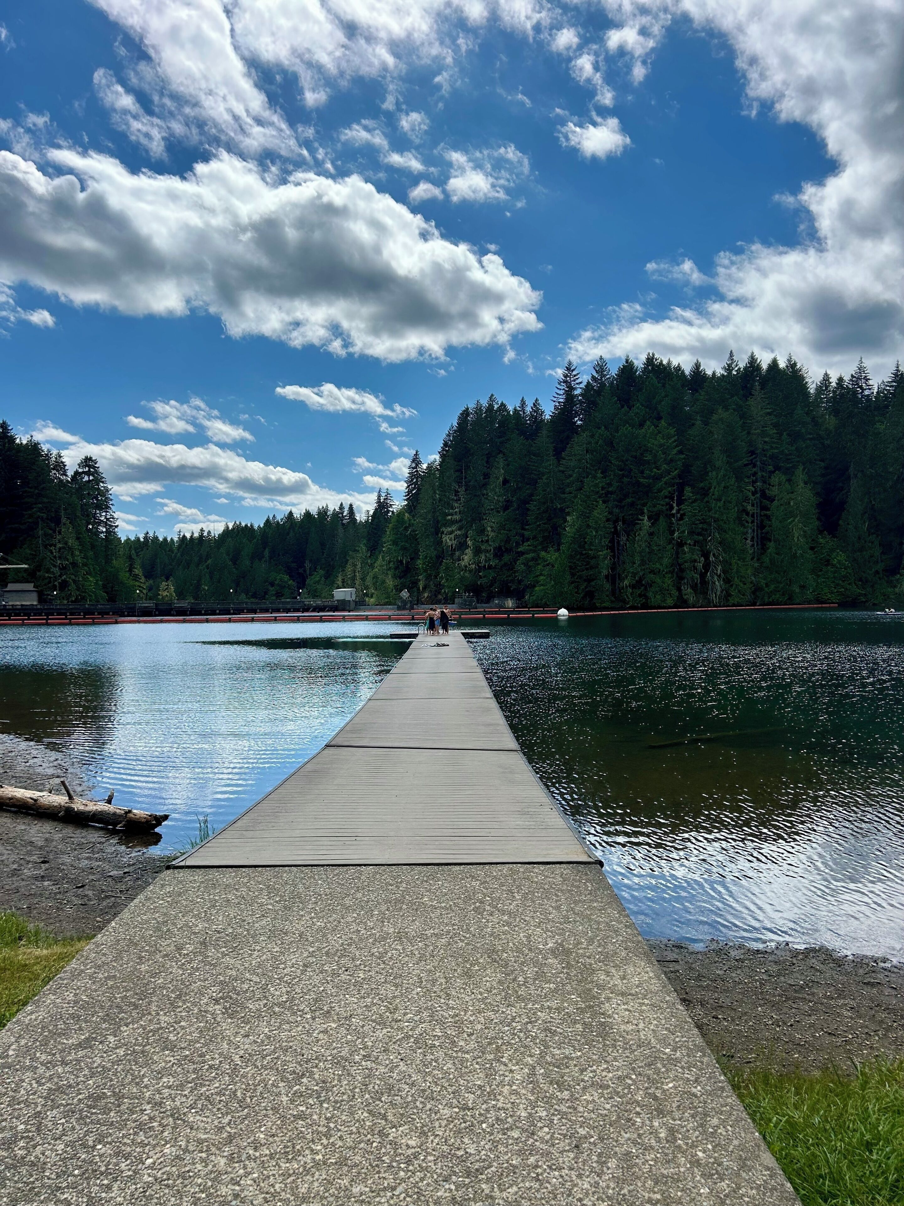 A concrete pier extending into a lake with a forested shoreline and a partly cloudy sky.