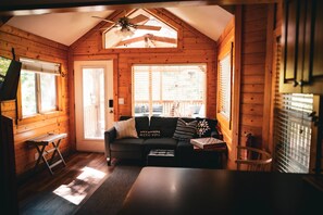 A cozy cabin interior with a ceiling fan, a black sofa with patterned cushions, a small table, and a window with blinds.