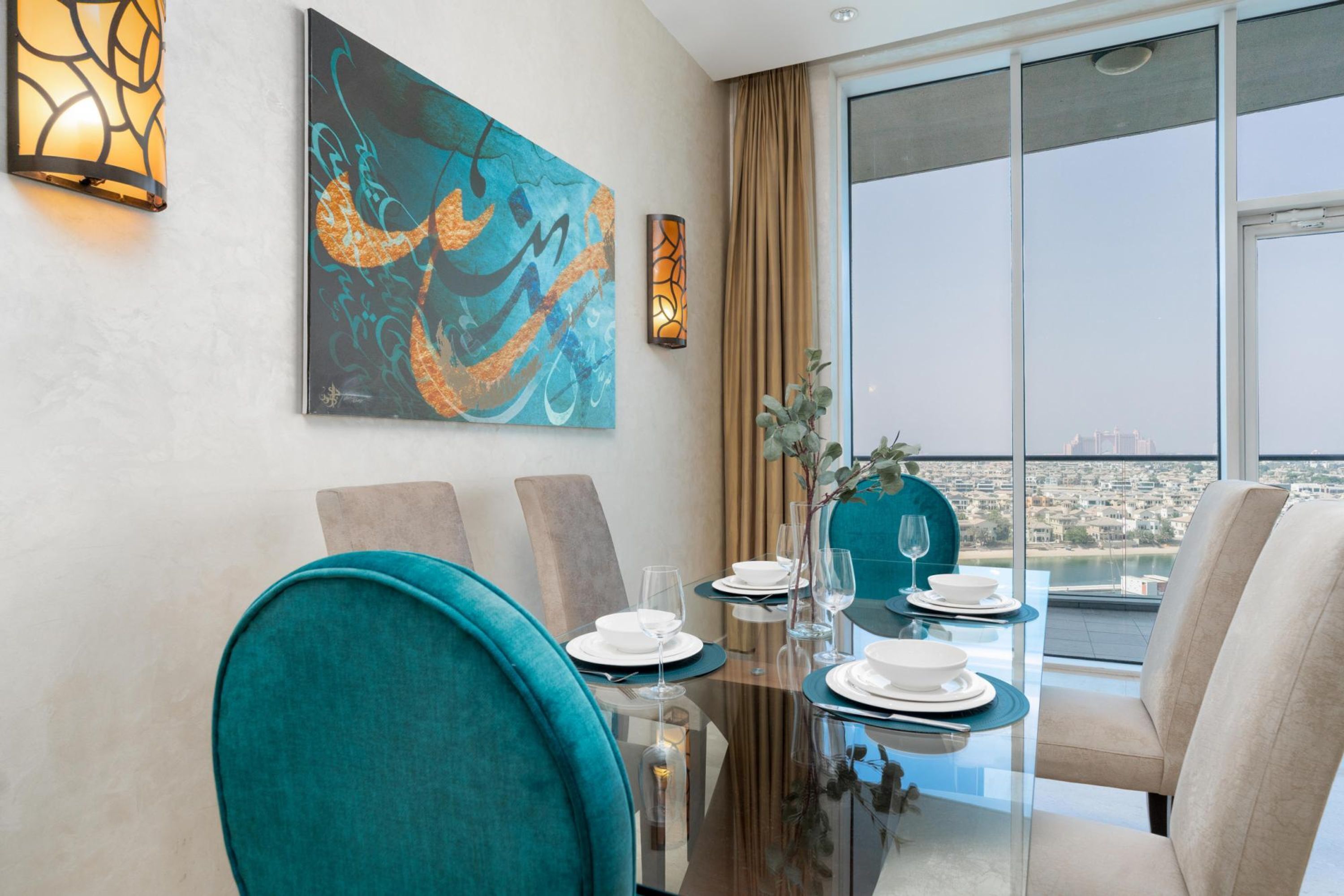 A dining area with a glass table, teal chairs, and a city view through large windows.