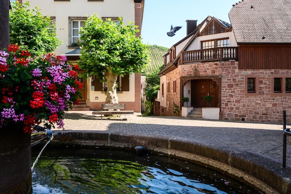 Une cour avec une fontaine, un banc et un bâtiment doté d’une façade en pierre et d’un balcon en bois.