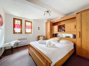 Un dormitorio con cama, armario de madera, dos ventanas con cortinas, una silla y un radiador.
