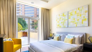 Ein Hotelzimmer mit einem großen Bett, einem gelben Sessel, einem Schreibtisch mit Lampe und Blick ins Freie durch eine Schiebetür.