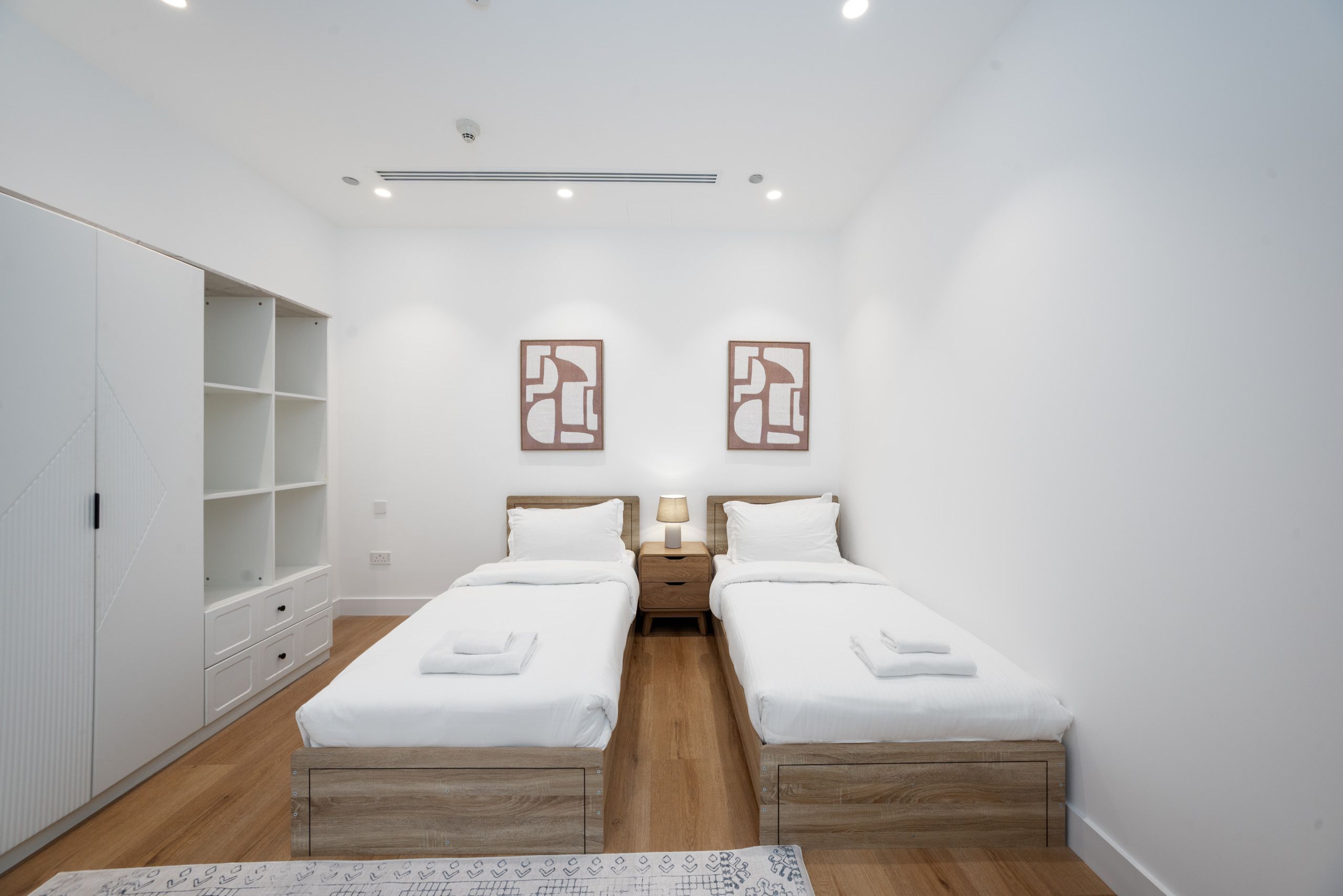 A modern bedroom with two single beds, a wardrobe, and a small bedside table with a lamp.