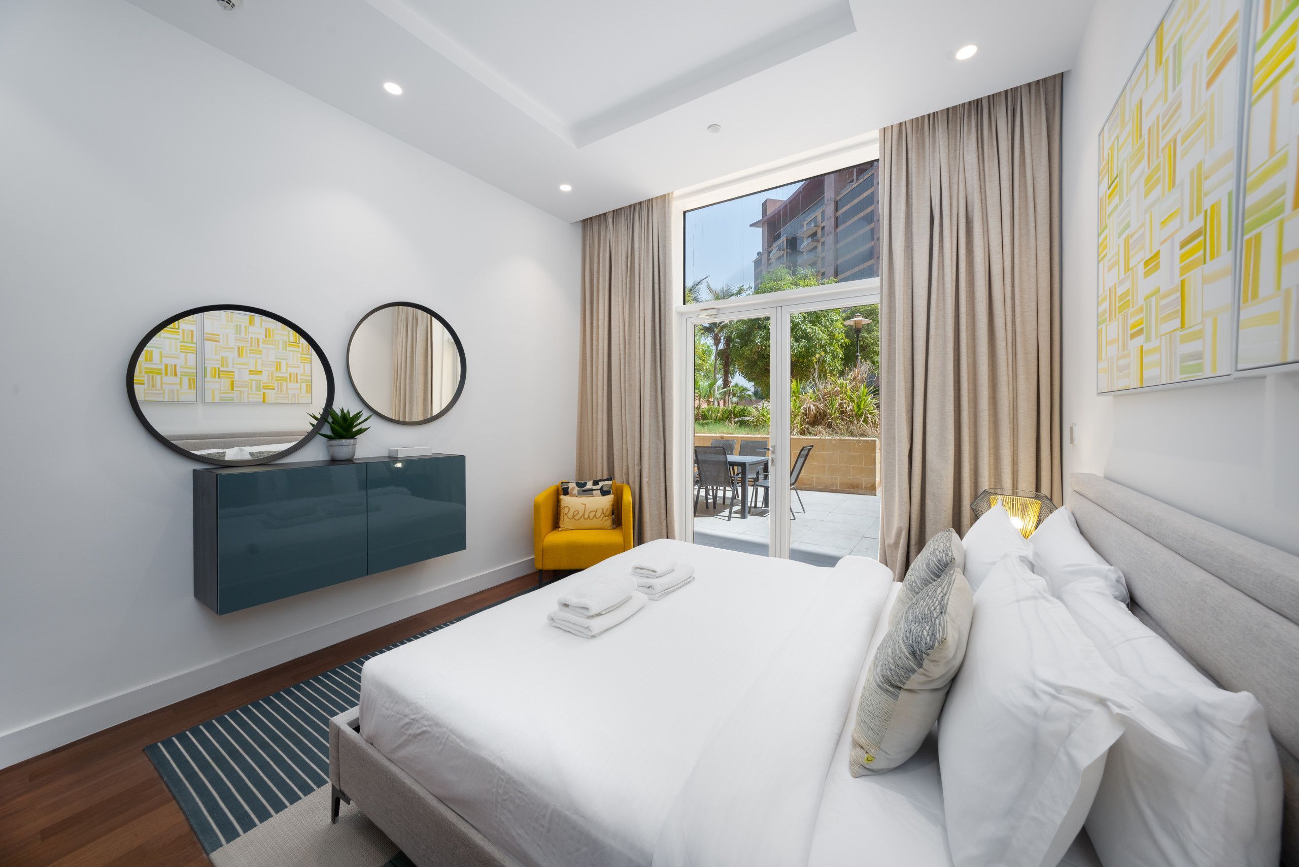 A modern hotel room with a bed, a vanity with a mirror, a small yellow chair, and a view of the outdoors through a large window.