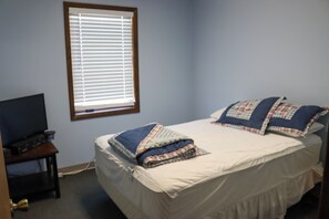 A bedroom with a bed, a window with blinds, a television on a stand, and a door with a brass doorknob.