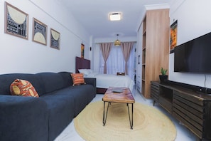 Uma sala compacta com sofá, mesa pequena, cama, televisão e obras de arte nas paredes.