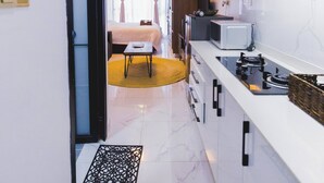 Un couloir étroit mène à une chambre comprenant un lit, une kitchenette équipée d’un four à micro-ondes et une petite table.