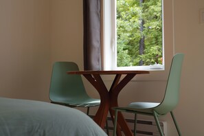 Una habitación con cama, una mesa con dos sillas, una ventana con vistas a árboles y un calefactor.