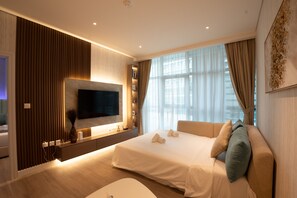 Una habitación de hotel moderna con una cama grande, un televisor de pantalla plana en la pared, una mesita de noche con un jarrón y una ventana con cortinas translúcidas.