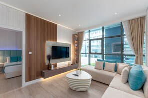 Una sala de estar moderna con un sofá, una mesa de centro redonda, un televisor de pantalla plana montado en un panel de madera y un amplio ventanal con cortinas.
