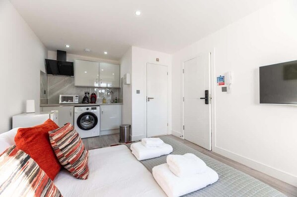 A compact hotel room with a bed, a washing machine, a kitchenette, and a TV mounted on the wall.