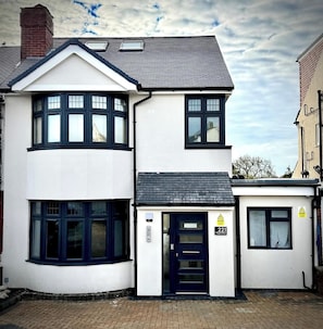 A two-story residential building with a dark blue front door, a small window with a yellow caution sign, and a brick pathway leading to the entrance.