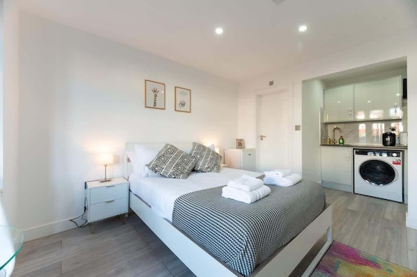 A modern hotel room with a bed, bedside tables, a washing machine, and a small kitchenette.