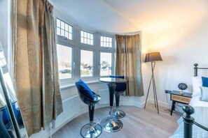 A room with a corner window, a round table, two bar stools, a floor lamp, a bedside table with a lamp, and a bed.