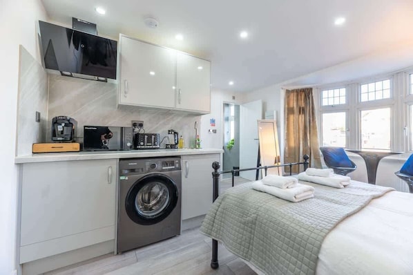 A compact hotel room with a bed, a washing machine, a kitchenette, a TV, and a window with curtains.