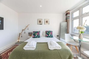 A hotel room with a bed, two pillows, a glass table, a chair, a TV, and a window with curtains.