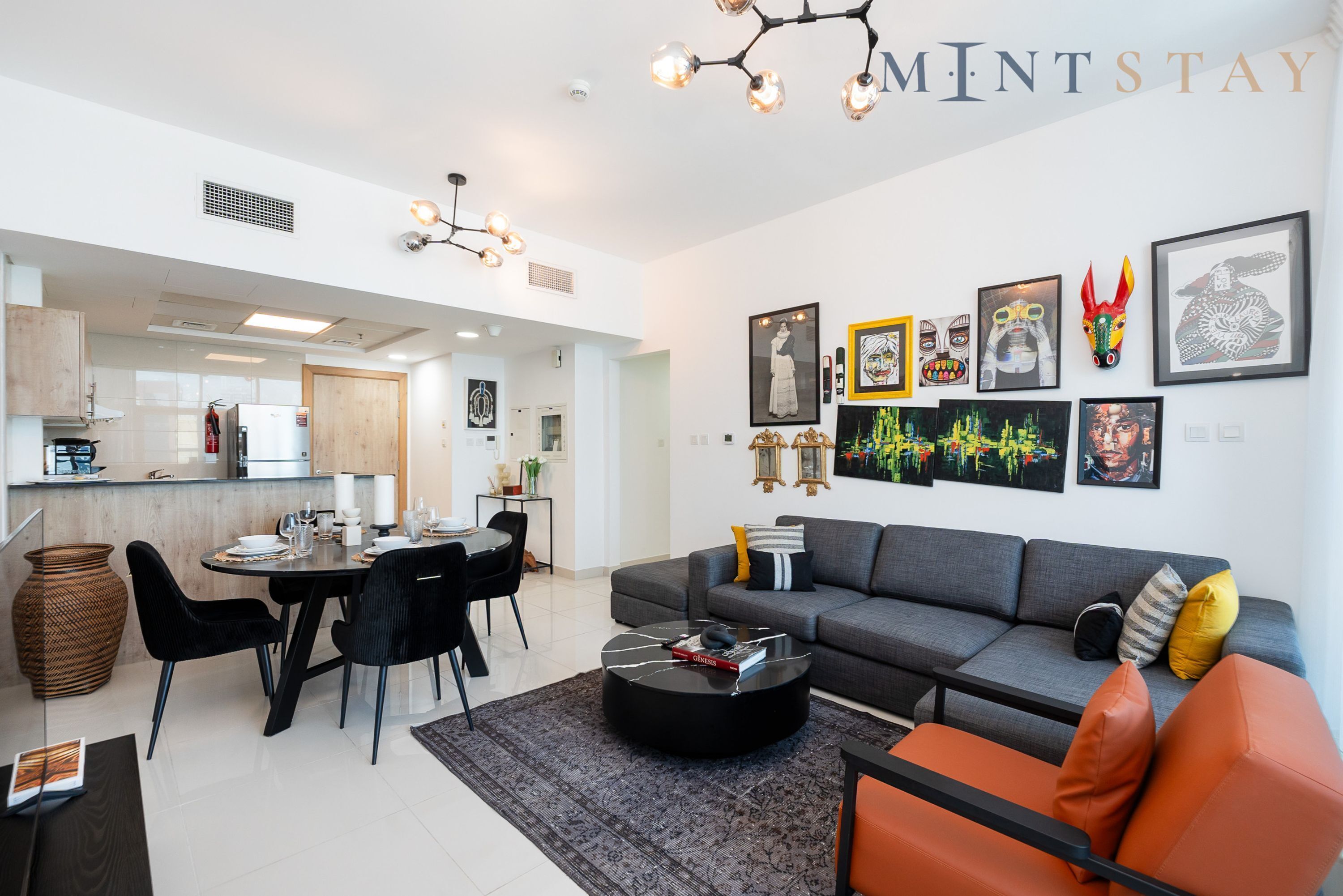 A modern living room with a grey sofa, black and orange chairs, a dining table, and a kitchen in the background.